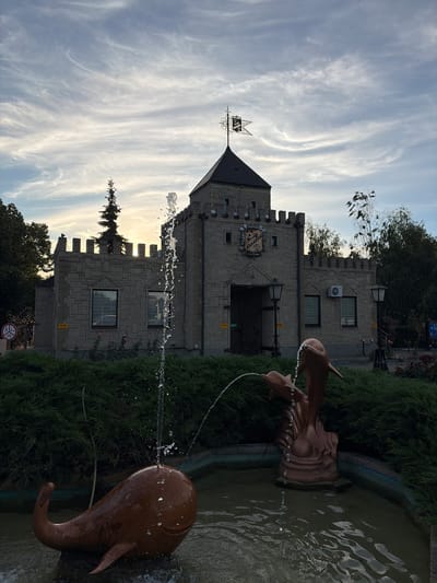 In the city of Slobozhanske, Ukraine, a stone building resembling a small castle stands under a cloudy sky. In the foreground, a fountain features a sculpture of a whale with water spraying from its tail and a group of dolphins with water arching from their snouts. Dense green bushes surround the fountain and the base of the building, with a few rose bushes visible near the entrance. The building has crenellated battlements, arched doorways, and windows with dark frames. A weather vane with a shield and flag is mounted on the main tower. The sky is filled with wispy clouds, suggesting either early morning or late afternoon light. No people are visible in the frame.