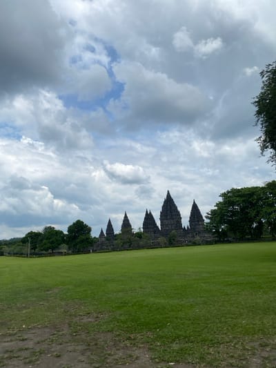 The image shows a view of the Prambanan temple complex in Kecamatan Prambanan, Indonesia, located at latitude -7.75, longitude 110.492. The scene is a wide shot of a grassy field with a large, grey stone temple complex in the background. The sky is cloudy with some blue patches visible. There are several trees in the foreground and the temple is visible in the distance. The grass is green and the ground is dirt. The temple is made of large, grey stones and has a distinct pyramidal shape. The image captures a panoramic view of the Prambanan temple complex, emphasizing its grandeur and historical significance amidst a serene landscape.