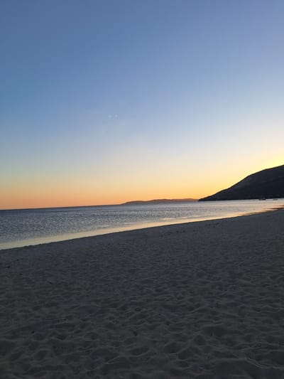 The image depicts a serene beach scene at dusk, with the sun setting over the horizon. The sky transitions from a deep blue to a soft orange and yellow hue, indicating the golden hour. A dark, silhouetted hill or mountain range can be seen in the distance, bordering the sea. The sea itself appears calm, with gentle ripples reflecting the soft light of the setting sun. To the right of the water, a sandy beach stretches out, with subtle undulations and textures in the sand suggesting the natural patterns left by the wind or tide. The overall atmosphere is peaceful and tranquil, evoking a sense of calm and natural beauty. There are no people or man-made objects prominently featured in the image, emphasizing the untouched and natural aspect of the landscape. The time of day is clearly evening, given the colors of the sky and the low angle of the sun, which is not directly visible but implied by the light. The location, Erdek, Turkey, is known for its beautiful coastline, and this image captures the essence of such a place. No text is visible in the image.