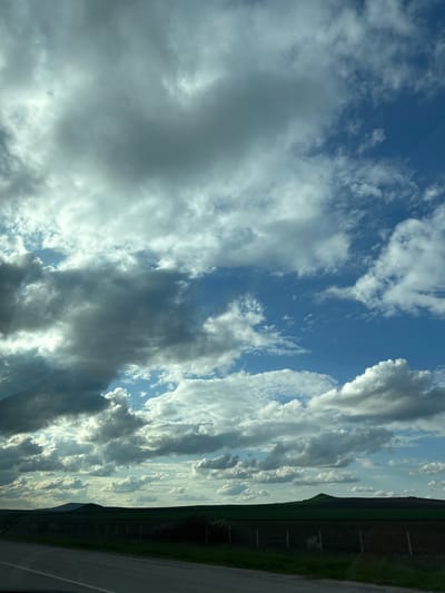 The image depicts a cloudy sky with visible patches of blue. The clouds are varied, ranging from fluffy white to dark grey storm clouds. In the upper right corner, there is a glimpse of land, likely a field and possibly some trees. A grey surface, probably a road, is visible at the very bottom right. The image is rotated 90 degrees counterclockwise. The location, Zlatina, Bulgaria, is not discernible from the visual content of the image.