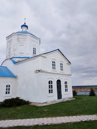 The image captures a white Orthodox church or chapel under an overcast sky. The building features a prominent blue dome topped with a cross, rising from an octagonal, tower-like structure with arched windows. Other sections of the church also have blue roofs and rectangular or arched windows with white frames. The white exterior walls show some signs of wear, particularly on the octagonal section, where paint is peeling. A dark wooden door is visible on the side of the main building.

In the foreground, a manicured green lawn surrounds the church, with a dark green bush situated near the left side of the structure. A patterned paved path, composed of white and reddish-brown square tiles, runs along the bottom edge of the frame. To the right, two small evergreen trees are planted on the grass, behind which a metal fence separates the church grounds from the background.

The background reveals a rural landscape of rolling, partially vegetated hills stretching towards the horizon, beneath the same grey, cloudy sky. Faint power lines or telephone wires are visible in the distance. The lighting suggests a dull, overcast daytime. No people or visible text are present, and the scene is static and quiet, depicting a religious building in a serene, semi-rural setting in Yablonovo, Russia.