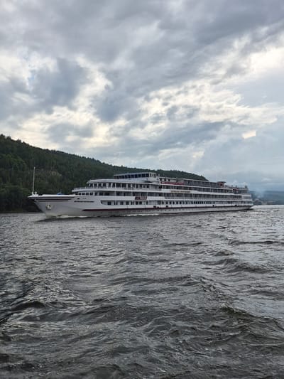 A large, white, multi-deck river cruise ship, named "Мстислав Ростропович" (Mstislav Rostropovich), sails on a dark, choppy river. The ship is the primary subject, positioned centrally and taking up a significant portion of the image's horizontal space. It has numerous windows along its multiple decks and a distinct red stripe along its waterline. The ship's bow is pointed slightly to the left, creating a dynamic sense of movement.
Behind the ship, on the left side of the frame, a densely forested, green hillside rises sharply from the riverbank. This appears to be the location of the village of Shiryaevo, Russia, known for its picturesque natural setting along the Volga River. The sky above is overcast with dramatic, textured gray clouds, suggesting a moody or approaching weather event. The water's surface is disturbed with numerous ripples and small waves, indicating wind or the passage of the ship. No people are clearly visible on the ship or in the surrounding environment. The overall scene is one of travel and nature, with the imposing cruise ship contrasting with the wild, forested landscape under a dramatic sky.