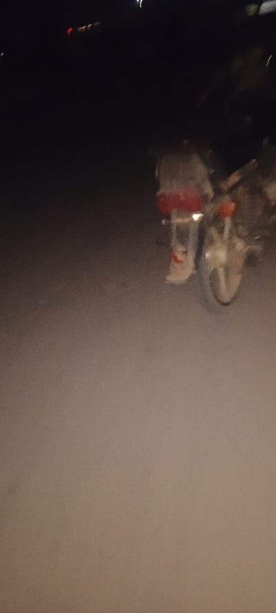 A blurred, low-lit image reveals a motorcycle parked on a paved surface, possibly a road or parking lot. The rear of the motorcycle is visible, featuring a red taillight and a partially visible tire. The overall darkness and lack of focus make details difficult to discern. There are lights in the upper left corner, but they are too blurry to make out anything.