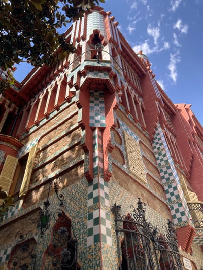 This is a low-angle, full-shot photograph of the exterior of Casa Vicens, located in the Gràcia district of Barcelona, Spain. The building exhibits a vibrant and eclectic architectural style, characterized by its red brick facade adorned with colorful ceramic tiles. These tiles feature intricate geometric patterns, checkerboard designs in green and white, and floral motifs in yellow and green. The building's upper sections are decorated with red brickwork and contrasting turquoise and white checkered tiles.
Several architectural elements are visible, including arched windows with red decorative framing, a small balcony with a wrought-iron railing, and elaborate wrought-iron gates at the bottom of the frame. A single green glass lantern hangs from a decorative wrought-iron bracket on the wall. The sunlight casts strong shadows across the textured stone portions of the building's lower facade, highlighting the details of the stonework and the tiled surfaces. A portion of a leafy tree branches in from the upper left corner of the image, providing a natural contrast to the man-made structures. The sky above is a clear blue with scattered white clouds. There are no people visible in the image.