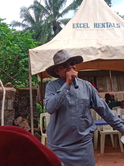 An elderly man in Udi, Nigeria, speaks into a microphone while standing under a tent labeled "Excel Rentals." He is wearing a gray suit, a cowboy hat, and sunglasses. People sit in white plastic chairs behind him. There is a brick wall and foliage in the background.