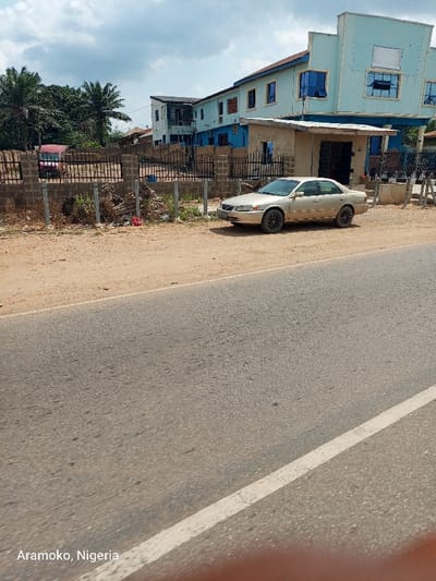 The image shows a light-colored sedan parked on the side of a paved road. The car is facing the camera and is parked next to a black metal fence. Behind the fence is a two-story blue building with a white roof. There are trees and vegetation visible in the background. The road is lined with a white stripe. The scene is located in Aramoko, Nigeria (latitude: 7.832, longitude: 4.515).