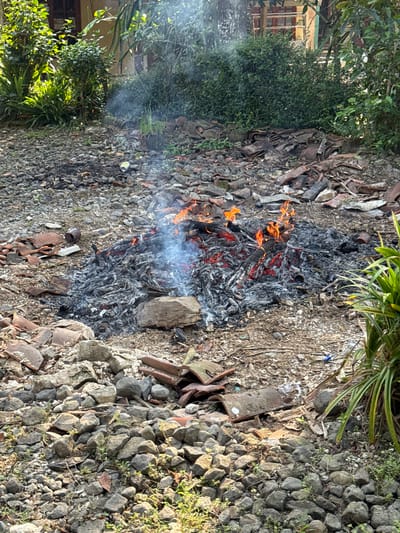 The image depicts an outdoor scene in Kalirancang, Indonesia, likely a backyard or a less-maintained area. In the foreground and midground, a pile of smoldering ashes and burning debris is the central focus. Flames are visible, indicating active burning, and smoke is rising from the pile. The debris appears to consist of dry leaves, twigs, and possibly remnants of old roofing tiles, as broken terracotta-colored pieces are scattered around the burning pile. The ground is covered with a mixture of gravel, rocks of various sizes, and dirt. Some larger rocks are also present, particularly in the lower half of the image.
In the background, lush green foliage and vegetation are visible, suggesting the presence of trees and bushes. A building with wooden window frames and what appears to be a tiled roof is partially visible behind the greenery. The overall impression is one of a somewhat untidy outdoor space where waste is being cleared by burning. No people are present in the frame. The lighting suggests it is daytime, with sunlight casting shadows.