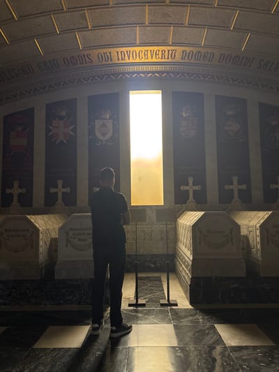 A man stands in the Royal Pantheon of San Lorenzo de El Escorial, Spain, facing a tall, narrow window that emits bright, diffused light. The man, silhouetted against the light, is viewed from behind, wearing a dark t-shirt and dark pants, with white-soled athletic shoes. He has a backpack on. The Pantheon is dimly lit, with the man's presence being the primary human element.

The space features a vaulted ceiling with visible rectangular panels. A decorative golden inscription runs along the upper wall, with the visible text reading "IS: ET GRIT OMNIS QUI INVOCAVERIT NOMEN DOMINI SALV" and continuing further along the curve. Below this, a decorative border of interlocking X shapes is present.

Flanking the window are several dark rectangular panels, each adorned with a coat of arms topped by a crown. Below these are large white crosses. These are set above rows of sarcophagi, which are ornate and carved, with inscriptions. One sarcophagus visible on the left has an inscription that appears to read "Similis factus est leoni in operibus suis." Another in the center reads "Non moriar sed vivam et narrabo opera Domini." A third on the right has the inscription "Vita ex morte." The sarcophagi are made of white marble and are placed on a dark, polished marble floor with a checkerboard pattern of light and dark squares. The walls behind the sarcophagi are also of dark, polished marble. Two tall, thin black poles with square bases are positioned in front of the sarcophagi, likely acting as barriers or for display.