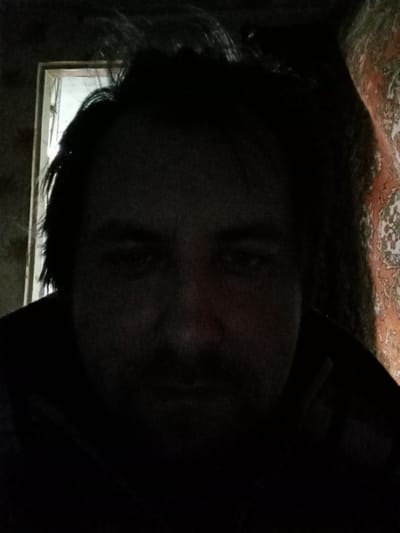 A dimly lit selfie of a man with dark hair and a beard. The background is indistinct, but appears to be an interior space with a doorway or window to the left and a patterned wall covering to the right. Due to the low lighting, specific details of the man's face and clothing are difficult to discern. There is no information that can be used to confirm if this image was taken in Parabel, Russia.