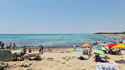 The image depicts a crowded beach scene in Porto Cesareo, Italy, on a clear, sunny day. The turquoise water of the Ionian Sea stretches to the horizon, with a few small boats visible in the distance. The sandy beach is populated with numerous people, many of whom are swimming, sunbathing, or relaxing under colorful umbrellas. Several beach umbrellas are set up, providing shade for beachgoers. On the shore, people are engaged in various activities, including walking, playing, and carrying beach gear. The foreground shows a rocky area with sand, and further back, the beach extends to a line of trees and vegetation. The overall atmosphere is one of a vibrant summer day at the beach.
