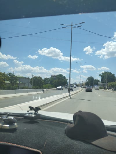The image shows a view from inside a car looking forward on a sunny day. The road is a multi-lane asphalt road with concrete barriers separating lanes. Several cars are visible on the road, including a silver sedan, a white minivan, and a dark sedan in the foreground. Streetlights are positioned along the road, and there are trees and buildings visible on either side. The sky is blue with scattered white clouds. The foreground of the image shows the dashboard of the car, with a small black device with suction cups and a dark baseball cap. Based on the visible streetlights and road infrastructure, this scene is consistent with the urban environment of Forish QFY, Uzbekistan.