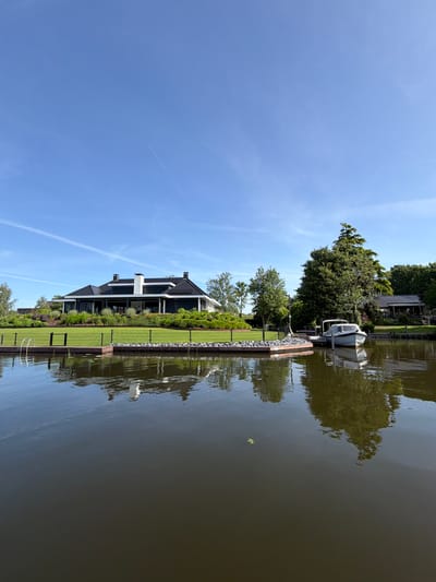 The image shows a waterfront property in Leerdam, Netherlands. A modern two-story house with dark trim sits on a manicured lawn extending to the water's edge. A black fence separates the lawn from the waterway. A small boat is docked near a pier, and trees line the opposite bank. The water is calm, reflecting the house and surrounding vegetation. The sky is clear blue with faint contrails.