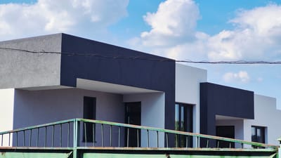 The image depicts a modern, multi-story building with a contemporary architectural design. The building features contrasting colors, with sections painted in dark gray and white. There are visible windows and overhangs. In the foreground, a green metal fence with vertical bars runs horizontally across the lower portion of the image. Overhead, a series of thin black wires are suspended against a bright blue sky with scattered white clouds. The scene appears to be an exterior view of a residential or commercial property. The metadata indicates the location is Ialoveni, Moldova. No people are visible in the image.