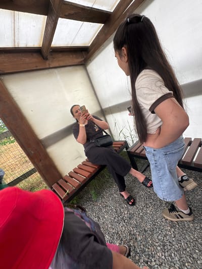 In the city of Zaliznychne, Ukraine, the image captures a scene under a covered structure with a wooden frame and translucent roofing. A woman is seated on a wooden bench, holding a mobile phone and appearing to take a picture. Another young girl, facing away from the camera, stands nearby, wearing a light-colored t-shirt and denim shorts. The ground is covered with gravel. In the foreground, partially visible, are the heads of two individuals, one wearing a red hat. The background shows a fence and vegetation.