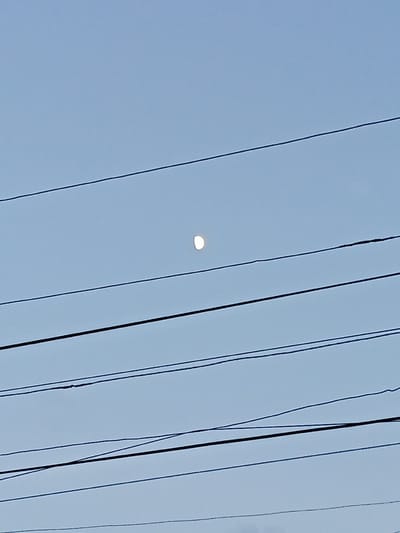 The image depicts a clear, pale blue sky with several horizontal black power lines crisscrossing the frame. In the upper-middle section of the image, a partially illuminated moon is visible. The moon appears bright white with a slight crescent shape. The power lines are of varying thickness and are positioned at different heights, creating a layered effect against the sky. The scene is devoid of any people or discernible landmarks, focusing solely on the sky, the moon, and the man-made electrical infrastructure. The lighting suggests it is daytime, possibly late afternoon or early morning, given the brightness of the sky and the visibility of the moon. There is no visible text within the image. The overall impression is a simple, almost minimalist composition highlighting the contrast between the natural celestial body and the man-made power lines in a sky that suggests a calm, clear atmosphere.