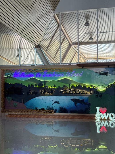 This image shows a brightly lit, colorful mural depicting a landscape and marine life, with the words "Welcome to Sumbawa Island" written in neon lights at the top. The mural features a stylized depiction of hills, a town with minarets, a bridge, and a plane in the sky. The foreground of the mural shows a body of water with coral, fish, a diver, and a whale shark. On the right side of the mural, the words "I LOVE SOWQ" are displayed in large, illuminated letters, with the "O" in "LOVE" replaced by a heart symbol that is lit red. The mural is reflected on a polished floor below. The background of the image shows a modern ceiling with a slatted design and suspended lights, suggesting an indoor public space, possibly an airport terminal or a visitor center, in Lempeh, Indonesia.