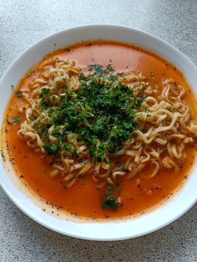 A close-up, overhead shot shows a white bowl filled with ramen noodles in a red broth, garnished with chopped parsley. The noodles are thin and wavy, submerged in a vibrant orange-red broth. Dark flecks, likely spices or herbs, are visible throughout the broth and around the rim of the bowl. A generous portion of fresh, bright green parsley is piled on top of the noodles, adding a fresh contrast to the warm colors of the dish. The bowl rests on a speckled gray surface, possibly a countertop or table. The lighting suggests it is daytime, and the overall impression is one of a simple, comforting meal. There is no visible text in the image.