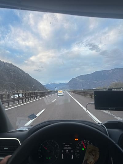 The image depicts a first-person perspective from inside a vehicle traveling northbound on Autostrada A22, specifically within the Adige Valley near Vadena - Pfatten, Italy. The scene is observed through the vehicle's windshield, which exhibits minor smudges.

In the immediate foreground, the top portion of a black steering wheel is visible, along with parts of the dashboard. The instrument cluster shows the speedometer indicating approximately 60 km/h, the tachometer, and a digital display showing "Check AdBlue" and "Pollution System." A yellow warning light is illuminated. A small, light-colored decorative object is affixed to the dashboard near the speedometer. To the right of the steering wheel, a black monitor, branded "VIVAL," is mounted on the dashboard, connected by a cable. A partial view of a dark-skinned hand is visible on the lower left of the frame, gripping the steering wheel.

The highway ahead consists of at least two lanes in the direction of travel, separated by solid and dashed white lines, with a wet asphalt surface suggesting recent precipitation. In the middle lane, a white utility van with yellow markings, including a partially visible "HO" on its rear, is traveling ahead of the viewer's vehicle. To the left, a concrete barrier separates the opposing lanes of traffic, where several silver-colored cars are visible traveling southbound. A brown metal guardrail runs along the right shoulder of the highway.

The environment is characterized by mountainous terrain under an overcast sky with broken clouds. Steep, forested mountains flank both sides of the valley. In the distant background, snow-capped peaks are visible. A bridge structure with several pillars is seen to the far left, beyond the opposing traffic lanes. The overall atmosphere is cool and damp.