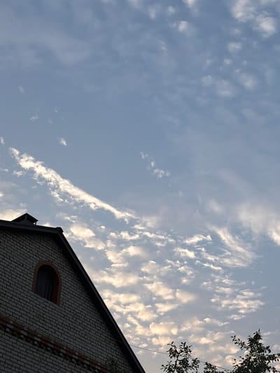 The image shows the roof and upper portion of a brick building against a sky filled with wispy white clouds. The building has a dark roof with a visible chimney. A distinct arched window is visible on the side of the building, which is constructed with light-colored bricks and features red brick accents. The sky is a pale blue, with sunlight illuminating the clouds from behind, giving them a soft, glowing appearance. The bottom right corner of the image shows the dark leaves of trees. The scene appears to be captured during daylight, possibly in the late afternoon or early evening, given the lighting conditions. No people are visible in the image. The location is identified as Leninsk-Kuznetskiy, Russia.