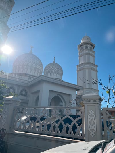 This image, taken in the city of Lambaro, Indonesia, features a white mosque with multiple domes and a tall minaret. The mosque's architecture is detailed with intricate patterns on the domes and ornate window designs, one of which displays Arabic calligraphy that appears to be the word "Allah." In the foreground, a white decorative fence with geometric cutouts separates the viewer from the mosque. Behind the fence, the tops of cars are visible, suggesting a street or parking area. The sky is a clear, bright blue, and the sun is shining intensely, causing lens flare in the upper left portion of the image. Power lines stretch across the top of the frame, and a few bare tree branches are visible in front of the minaret and to the right. The overall scene is one of a bright, sunny day in an urban or suburban setting, highlighting the mosque's prominent presence.
