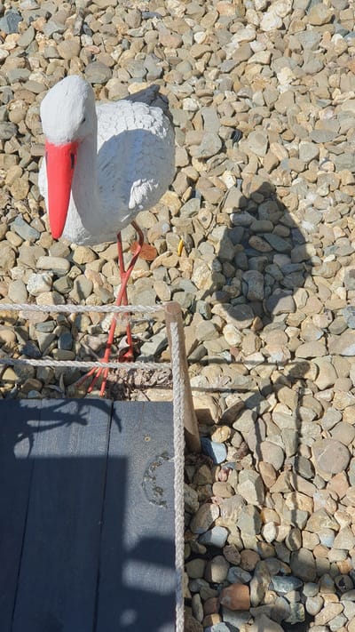 The image shows a concrete statue of a stork with a red beak and red legs, standing on a gravel surface. The stork statue is white with black markings on its wings. The background consists of various sized stones and pebbles. In the foreground, there is a dark wooden plank and a rope barrier. The scene appears to be outdoors. There are no people or actions visible in the image. The location is identified as being in the city of Ust-Labinsk, Russia.