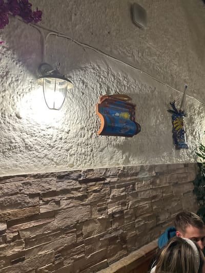 The image depicts an outdoor, nighttime scene, showcasing two distinct wall textures. On the left and center, a rough, white stucco wall is illuminated by a bright, vintage-style light fixture with a metal cage, casting light downwards. Mounted on this wall are two decorative, tile-like pieces: a larger, curved one featuring blue and gold patterns, including a sun and possibly marine life, and a smaller, rectangular piece with ribbons. A white utility box or speaker is also present in the upper left, with white electrical cables running along the wall.

To the right, a vertical section of a stone-clad wall with varying shades and sizes of rectangular stones extends upwards. In the upper right corner, the heads and shoulders of two individuals are partially visible. One person, with short dark hair, wears a blue garment and looks forward. Adjacent is another person with long brown hair and a sparkly headband, also looking in the same direction, suggesting an intimate interaction or shared focus. Dark green foliage is visible at the top, and a cluster of purple flowers appears in the bottom left. The overall setting, with its decorative elements and distinct architecture, evokes a Mediterranean atmosphere, consistent with the location context of Agios Dimitrios, Greece. There is no visible text.