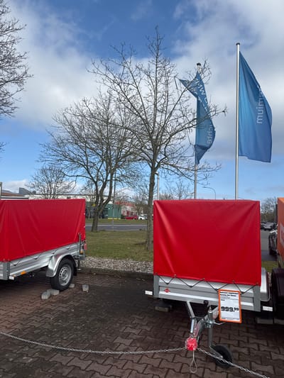 The image displays an outdoor retail display area under a partially cloudy sky, located in the retail park on Marburger Straße in Gießen, Germany, near an OBI hardware store and a BMW dealership.
In the foreground, two identical single-axle utility trailers with silver metal frames are parked on a dark grey paver-stone surface. Each trailer is fitted with a bright red, taut canvas cover. The trailer positioned to the right features a white price tag attached to its A-frame, displaying "999,99" Euros, preceded by the German text "Anhänger ungebremst Verdeck Set Sirius" (unbraked trailer canopy set Sirius). Small grey concrete blocks are visible on the ground near the wheels of the left trailer, serving as chocks or display stands. A silver chain stretches horizontally across the lower foreground, demarcating the display space.
The midground features several bare deciduous trees with dark, visible branches, indicating winter or early spring foliage conditions. Behind these trees, a paved road is visible. To the right of the trees, two tall flagpoles rise. The left flagpole holds a blue flag with white lettering, partially displaying "BMW", identifying a nearby BMW dealership. The right flagpole displays a blue flag with white lettering that reads "muim" (mixim spelled backward), a characteristic branding element for an OBI hardware store.
In the background, across the road, commercial buildings are present. These include a structure with a yellow-accented facade on the left and a red-roofed or red-sided building further to the right. Multiple vehicles, including a red car, are visible on the road and in distant parking areas. The sky is partially covered by white clouds, with patches of clear blue visible. No individuals are present in the scene.