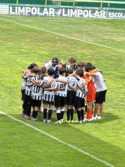 A group of young male soccer players, wearing black and white striped jerseys and black shorts, are huddled together in a circle on a bright green, well-maintained soccer field. Their arms are around each other's shoulders and backs. One player in the center is wearing an orange jersey and goalie gloves. A man in a white shirt and black cap, presumably a coach, is also part of the huddle, facing down towards the players. The background shows a green banner with white lettering that reads "LIMPÓLAR LIMPOLAR ESCOLA". The field is marked with white lines. The image appears to have been taken on a sunny day.