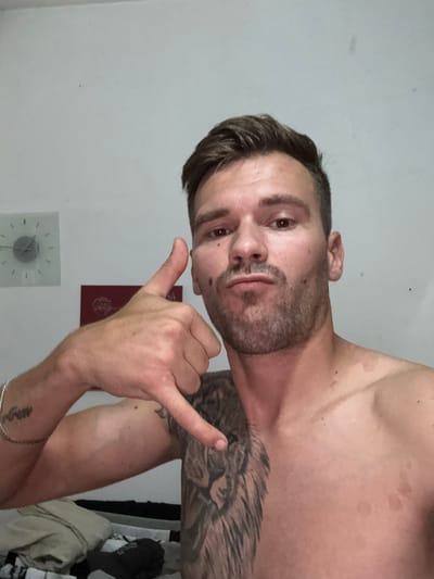 The image shows a man making a "call me" gesture with his right hand. He is shirtless and has a tattoo of a lion on his left shoulder and chest. He is wearing a bracelet on his left wrist. The background is a plain white wall with a clock on it. There are some reddish circular marks on his chest and shoulder. The man has short brown hair and a beard. He is looking at the camera with a neutral expression. The location is described as being in the city of Sesto Calende, Italy.