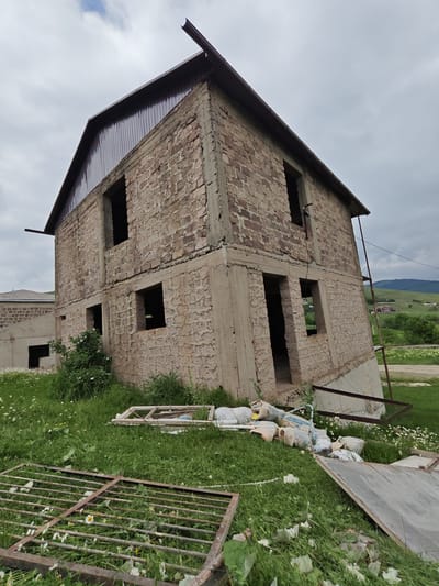 The image depicts an unfinished two-story building under a cloudy sky, suggesting it might be late afternoon or early evening. The building is constructed with brick and concrete, with visible rebar and scaffolding. The roof is partially covered with dark metal sheeting. There are open window frames and doorways, indicating construction is in progress.

In the foreground, there is a pile of construction materials, including bags of cement and what appears to be a discarded window frame. A rusty metal grate lies on the grass. The scene is set in a rural area, with rolling green hills and some distant buildings visible in the background, consistent with the provided location context of Gugark, Armenia. The overall impression is one of a construction site that has been temporarily abandoned, with a slightly overgrown and unkempt appearance due to the presence of grass and wildflowers around the building and materials.