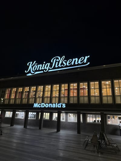 The image shows a McDonald's restaurant located in Duisburg, Germany, at night. A large neon sign above the restaurant reads "König Pilsener," and below it, another neon sign displays "McDonald's." The building has a dark brick facade with a row of illuminated windows on the upper level. In the foreground, there is a paved outdoor area with several chairs and tables, some of which appear to be made of wood. A person is visible on the far right of the image, standing near the building. The overall scene depicts an urban environment at night, with artificial lighting illuminating the signs and the interior of the restaurant.