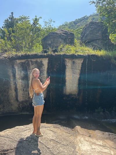 A young woman with long blonde hair is standing on a large rock, holding a pink smartphone and taking a photo. She is wearing a white bikini top and denim shorts. Behind her is a large, dark rock face with water trickling down. The scene appears to be a natural outdoor setting with lush green vegetation and trees on a hillside in the background. The sky is clear and blue.