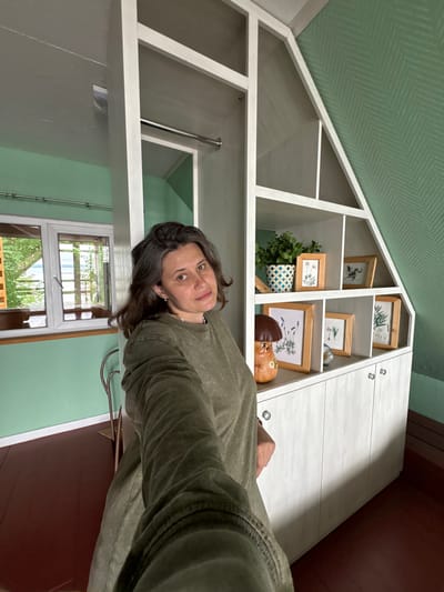 A woman with dark hair, wearing a green shirt, is taking a selfie in a room with green walls and a pitched ceiling. To her right is a white shelving unit with multiple compartments. The shelves contain decorative items including framed pictures of plants and birds, a plant in a patterned pot, and a ceramic mushroom figurine. Below the shelves are white cabinet doors with silver knobs. To the woman's left is a window with a view of greenery and water. The floor is dark wood.
