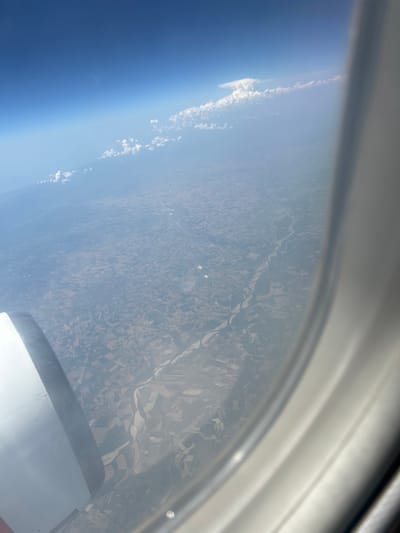 The image captures an aerial view from an airplane window, looking down on a landscape that features a meandering river cutting through agricultural fields. The scene is characterized by a hazy atmosphere, with patches of clouds visible in the distance against a bright blue sky. A portion of the airplane's engine is visible in the lower left corner, and the window frame occupies the right side of the image. The geographical location, identified as the area around Pavia, Italy, is suggested by the pattern of the river and the cultivated land. The fields are divided into geometric shapes, indicating a landscape shaped by agricultural practices. The river appears to be wide and shallow in some areas, with visible sandbanks or exposed riverbeds. The overall impression is one of a vast, rural landscape viewed from a significant altitude.