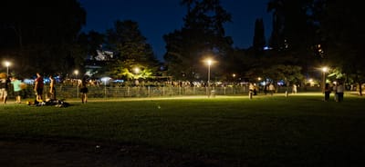 A nighttime park scene is depicted in Graz, Austria, likely an outdoor event or gathering. In the foreground, a well-lit grassy area is sparsely populated with a few people and some discarded items. A path runs along a fence separating the park from a more crowded area. Beyond the fence, numerous people are visible, some clustered around tents or stalls, suggesting a festival or market. Several lampposts illuminate the scene, casting pools of light on the grass and path. Trees and buildings are visible in the background, silhouetted against the dark sky. The overall impression is one of a lively evening event taking place in a park setting.
