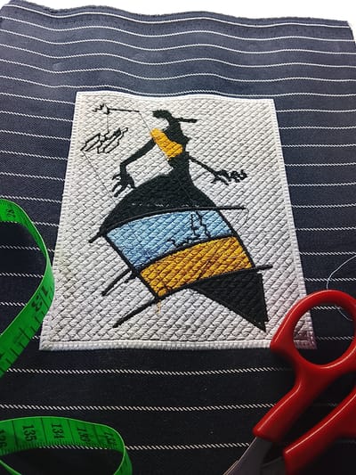 The image shows a close-up view of a piece of dark blue fabric with white pinstripes. On top of this fabric, there is an embroidered patch depicting a stylized silhouette of a person with a large, flowing dress. The dress is colored with sections of black, light blue, and yellow. To the left of the embroidered patch, a green measuring tape is visible, partially unrolled and curved. To the right of the patch, a pair of red-handled scissors is present. The overall scene suggests a crafting or sewing context. The image is not identifiable as being located in Rumuepirikom, Nigeria.