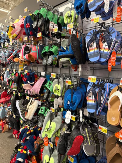 This image captures a vibrant and densely packed display of children's footwear within a retail store. The scene is dominated by multiple rows of shoes hanging from metal racks, suggesting an aisle dedicated to kids' summer and casual wear. No people are visible.

The collection features a wide variety of shoe types, including numerous pairs of flip-flops, many from the "Wonder Nation" brand, showcasing tropical beach sunset designs. Brightly colored mesh water shoes in blue, grey, and neon green are prominently displayed. Other notable footwear includes "Minecraft Creeper" themed slip-on sandals, character-themed slippers or sandals (such as a pink monster and a "Paw Patrol" design), and several pairs of athletic-style sneakers with varied soles, including one with a distinctive red tread.

Each pair of shoes is tagged with information. Yellow price tags clearly show prices such as "$5.48" and "$6.98". Orange tags often indicate the "Wonder Nation" brand and various children's shoe sizes, including "11-12" and "13-1". The footwear appears organized by type or size on the tiered hanging displays. The image is rotated, presenting the shoe racks vertically. The overall impression is one of an abundant and colorful selection of affordable children's shoes.