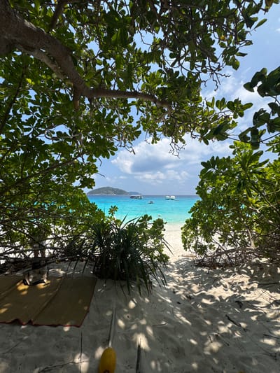 The image captures a view of a beach in Lam Kaen, Thailand. The perspective is from within foliage, with trees framing a path leading to the sand. The turquoise water is visible, with a few boats anchored offshore. The sand is light in color and appears to have been trodden on. To the right, a woven mat rests against the sand, with some light-colored object next to the mat.