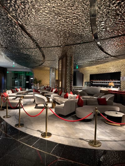 The image depicts a luxurious lounge area, likely a hotel lobby or a high-end bar, in Jianye District, China. The space is characterized by its opulent design, featuring a textured, metallic ceiling that reflects the ambient light. Plush, modular sofas in neutral tones are arranged in conversational groupings, accented with vibrant red throw pillows. Several round coffee tables are placed strategically between the seating areas.

The floor is a mix of polished dark stone and a light-colored carpet. Gold stanchions with red ropes delineate pathways and seating zones, adding to the sophisticated ambiance. In the background, a bar area is visible, with shelves displaying bottles and glassware, illuminated by warm lighting. The walls are decorated with textured panels and a series of framed art or decorative elements. The overall atmosphere is one of modern elegance and comfort. No people are visible in the frame.