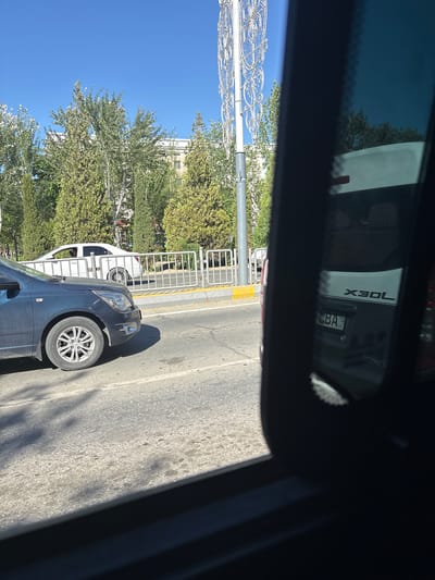 This media file captures a scene from inside a vehicle, looking outwards. The primary subject is the exterior of a dark-colored car parked on a paved road, with a white car visible behind it. The dark car's front wheel and fender are prominently featured, along with a portion of its windshield and hood. The background reveals a street lined with trees and a building, suggesting an urban or suburban environment. A metal fence or railing runs along the side of the road, separating the street from a green, tree-covered area. The sky is clear and blue, indicating daylight.

In the reflection of the vehicle's interior, possibly a windshield or side mirror, text is visible on the back of another vehicle. The text reads "X30L" and what appears to be a license plate with "LBA" on it. The lighting suggests it is daytime, likely late morning or early afternoon given the bright sunlight and distinct shadows. The scene appears to be a normal day, with no specific event or activity clearly depicted beyond vehicles being parked. The overall impression is a candid shot taken while stationary or moving slowly, offering a glimpse of the surroundings and the vehicle's reflection. The location context of Nukus, Uzbekistan, provides a geographical reference for the setting.