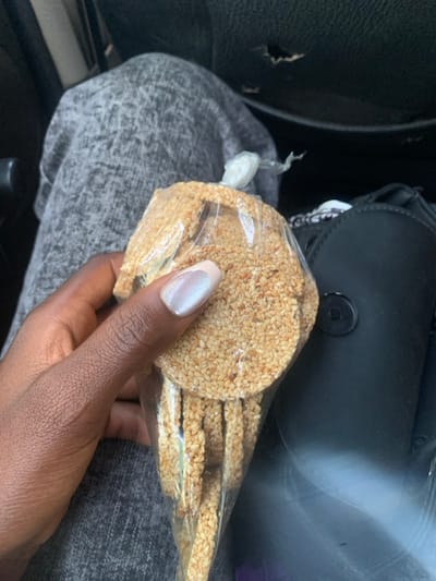 A dark-skinned left hand, featuring manicured nails with an iridescent light-colored polish and lighter tips, holds a transparent plastic bag. The bag contains multiple circular, flat snacks densely coated in sesame seeds, and is sealed at the top with a clear plastic knot. In the background, to the left, a portion of a leg clad in textured light grey fabric trousers is visible. To the right, a black object, consistent with a car seat or vehicle interior panel, shows a tear in its upper section and features a circular black clasp or button. The scene is situated within a vehicle in Rimin Ziem, Nigeria.