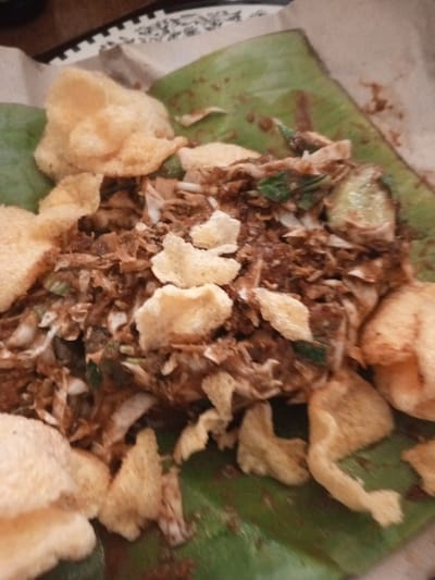 A plate of gado-gado, a traditional Indonesian salad, is presented on a banana leaf in Mvera Mission, Malawi. The dish consists of blanched vegetables, tofu, tempeh, and hard-boiled eggs, all coated in a peanut sauce. It is topped with krupuk, a type of cracker, and served with a side of lontong, compressed rice cake. The food is presented in a casual setting, likely a local eatery or a home-cooked meal.