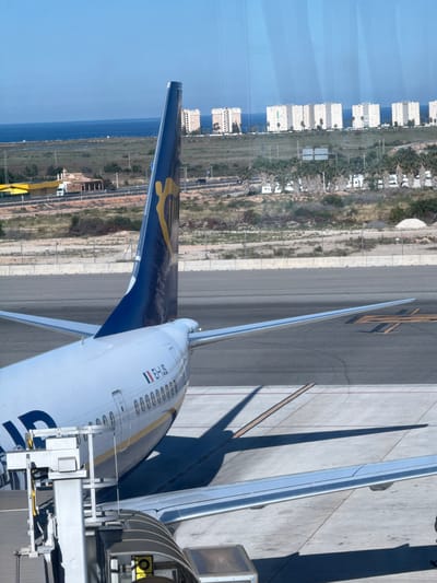 The image depicts the wing and fuselage of an airplane, likely at Alicante-Elche Miguel Hernández Airport near Elx/Elche, Spain. The plane is connected to a boarding bridge. The tail wing is visible with a blue and gold design. The fuselage is white with "EF-US" and a Spanish flag visible. Beyond the airport infrastructure, the landscape shows vegetation, buildings (possibly apartments or hotels), and a glimpse of the Mediterranean Sea in the distance. The scene appears to be taken during the daytime.