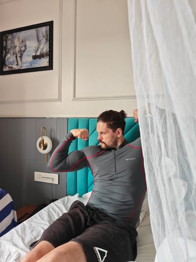 A man with his hair tied in a bun is sitting on a bed in a room, flexing his bicep. He is wearing a grey long-sleeved shirt with red trim and dark shorts. A white mosquito net hangs over the bed, partially obscuring the man. In the background, a framed picture of a white tiger in the snow hangs on the wall, and a bedside lamp is visible. The room appears to be a hotel room in Ajivali, India.