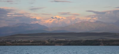 A wide, horizontal shot captures a tranquil scene of a lake in the foreground, leading to rolling hills and majestic mountains in the background. The sky above is a canvas of pastel hues, with soft pink and orange clouds scattered across a pale blue expanse. The mountains, tinged with the warm glow of sunset, are layered in varying shades of purple and blue, creating a sense of depth. The water in the lake is a muted blue, with subtle ripples disturbing its surface. The overall impression is one of serene natural beauty, possibly at dawn or dusk, in a mountainous region. The location is identified as Pristan'-Przheval'sk, Kyrgyzstan, a city situated on the eastern shore of Lake Issyk-Kul.
