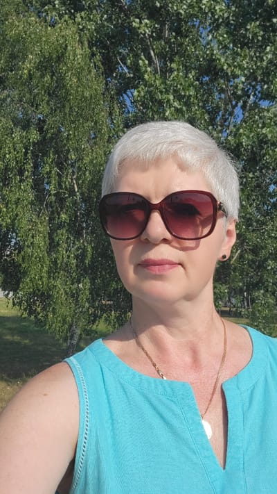 In the city of Belaazyorsk, Belarus, a middle-aged woman with short, grey hair and sunglasses stands outdoors, facing the camera. She is wearing a turquoise sleeveless top and a gold necklace. The background consists of lush green trees under a bright blue sky, suggesting a sunny day in a park or natural setting.