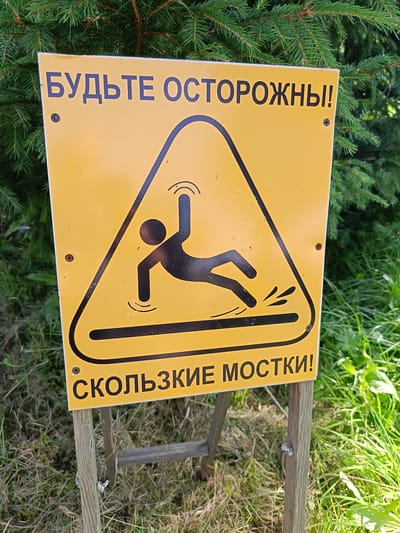 A yellow sign with a black border and text in Russian is mounted on two wooden posts in an outdoor environment. The sign features a black pictogram of a person slipping on a wet surface, indicated by water splashes. The text at the top reads "БУДЬТЕ ОСТОРОЖНЫ!" which translates to "BE CAREFUL!". The text at the bottom reads "СКОЛЬЗКИЕ МОСТКИ!" which translates to "SLIPPERY FOOTBRIDGES!". The background consists of green pine branches and green grass. The overall scene suggests a warning about slippery footbridges in Semenkovo, Russia.