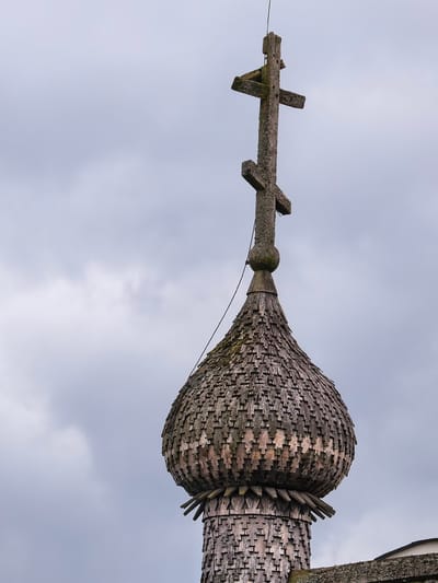 This is a close-up, eye-level, full shot of a wooden cupola topped with an Orthodox Christian cross, silhouetted against a cloudy sky. The cupola is covered in weathered, dark wood shingles that overlap to create a textured, bulbous shape. The cross is also made of aged wood, with a sloped lower bar and a smaller upper bar, giving it an octagonal appearance. A thin black wire extends from the cross down to the cupola, possibly for lightning protection. The overall aesthetic suggests an old, possibly historic, structure. The context provided specifies the location as Staraya Ladoga, Russia.