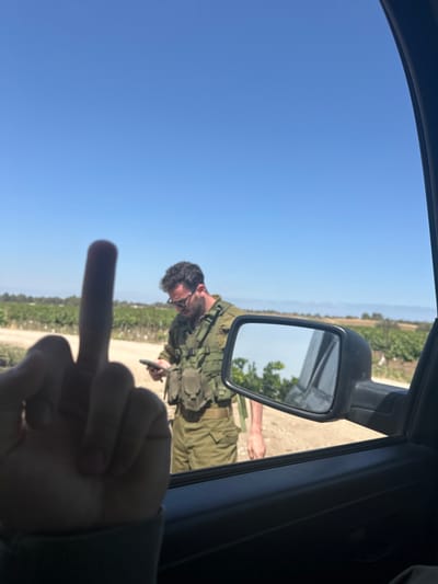 The image shows a person's middle finger raised towards a man wearing a military uniform in the background. The man in the military uniform is looking down at his phone. The scene appears to be taking place in a rural area with dirt roads and vegetation in the background. The location, based on the provided coordinates, is in Mazkeret Batya, Israel. The image is taken through the car window.