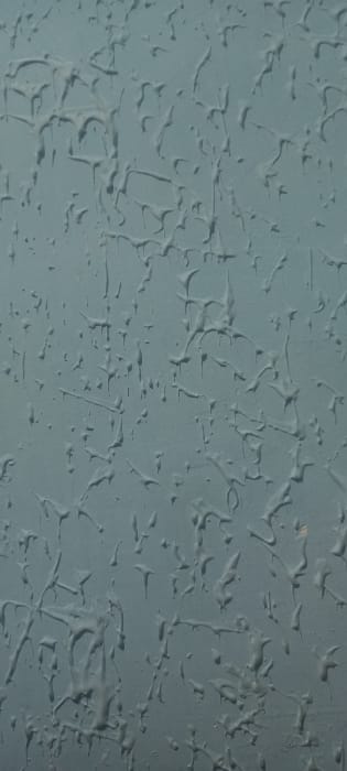 The image displays a close-up, vertical view of a textured surface, uniformly painted in a medium blue-grey color. The surface exhibits pronounced, irregular three-dimensional patterns resembling solidified drips, splatters, and elongated organic shapes. These raised textures vary in size and form, with some appearing as isolated teardrops or bumps, while others are interconnected, forming abstract networks across the plane. Consistent lighting, likely from an upper-left or overhead source, casts subtle shadows that define the relief and depth of these textured elements. No individuals, additional subjects, objects, or actions are visible within the frame. The visual content of the image provides no discernible features to confirm or identify the specific location as Kyzyltepa, Uzbekistan.